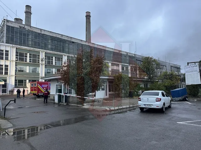 O clădire industrială lungă cu ferestre mari, un camion de pompieri roșu, un autoturism alb și un container albastru pe un teren asfaltat.