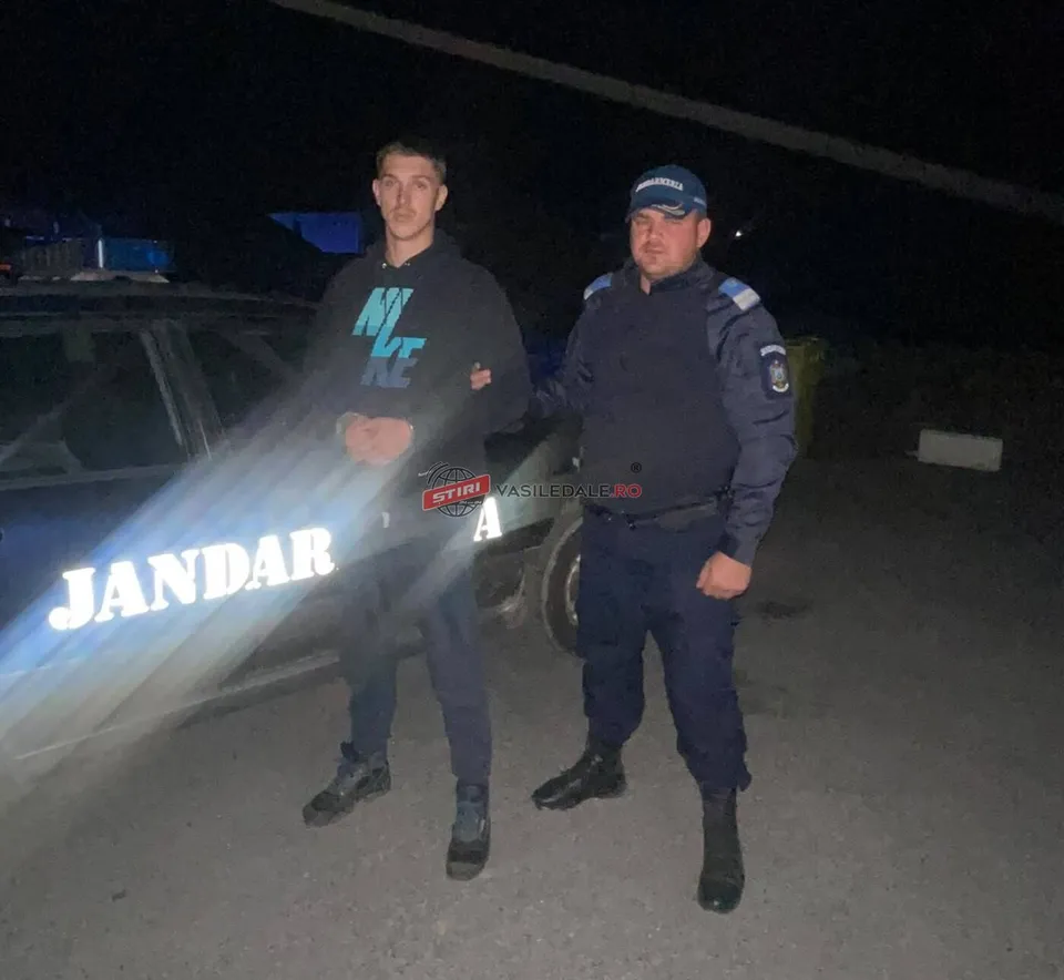 Un bărbat cu hanorac negru stă lângă un agent în uniformă albastră în fața unui vehicul cu inscripția JANDARMERIA.