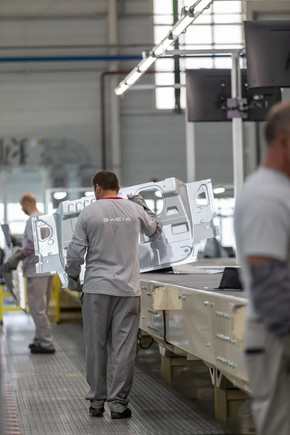 Un bărbat cu tricou gri și pantaloni de lucru gri manipulează o piesă metalică albă pe o linie de asamblare într-o fabrică.