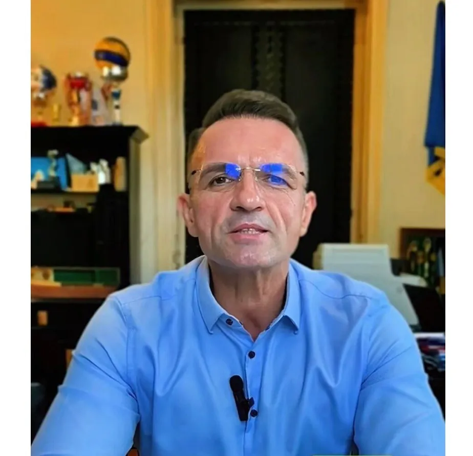 Un bărbat cu ochelari și cămașă albastră poartă un microfon de tip clip și privește spre cameră într-un interior cu un raft de trofee în spate.