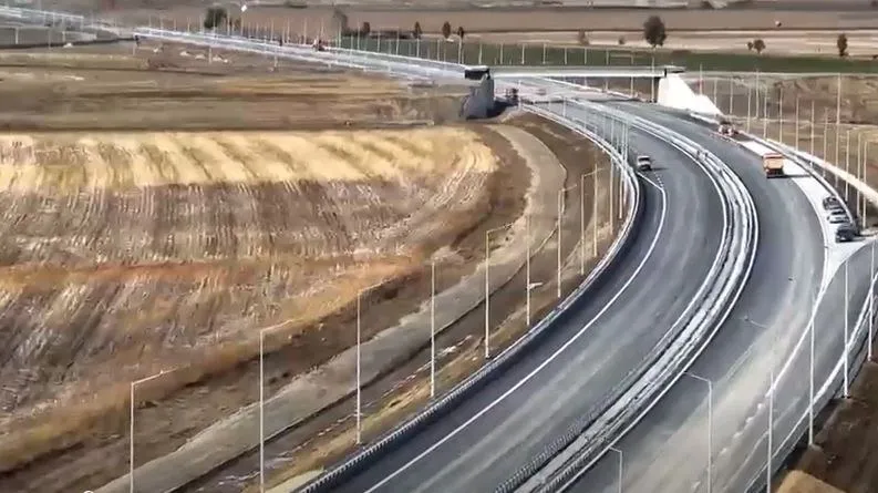 O porțiune de autostradă cu mai multe benzi de rulare curbe trece printr-un peisaj de câmp deschis sub un cer luminos.