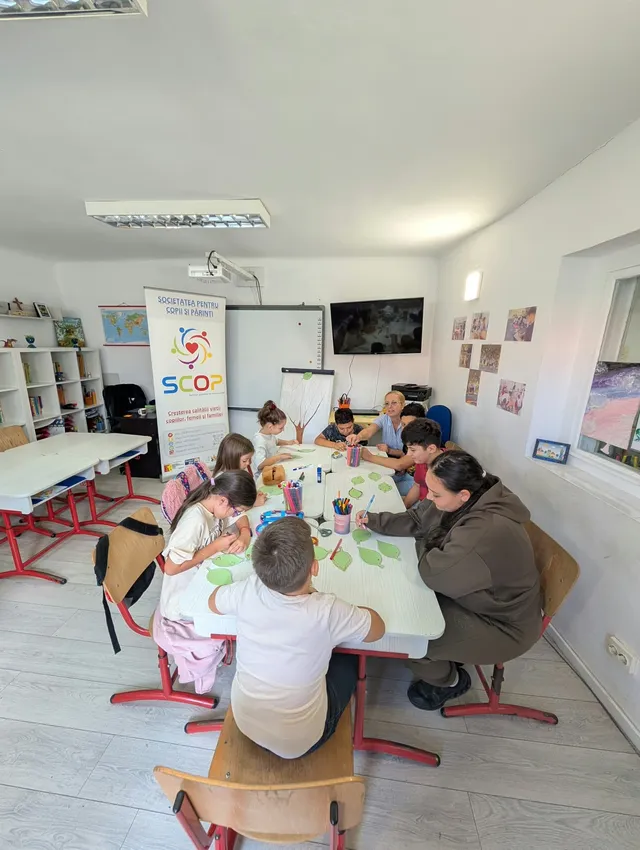 Un grup de copii și un adult stau la o masă albă și realizează activități de desen într-o încăpere luminată.
