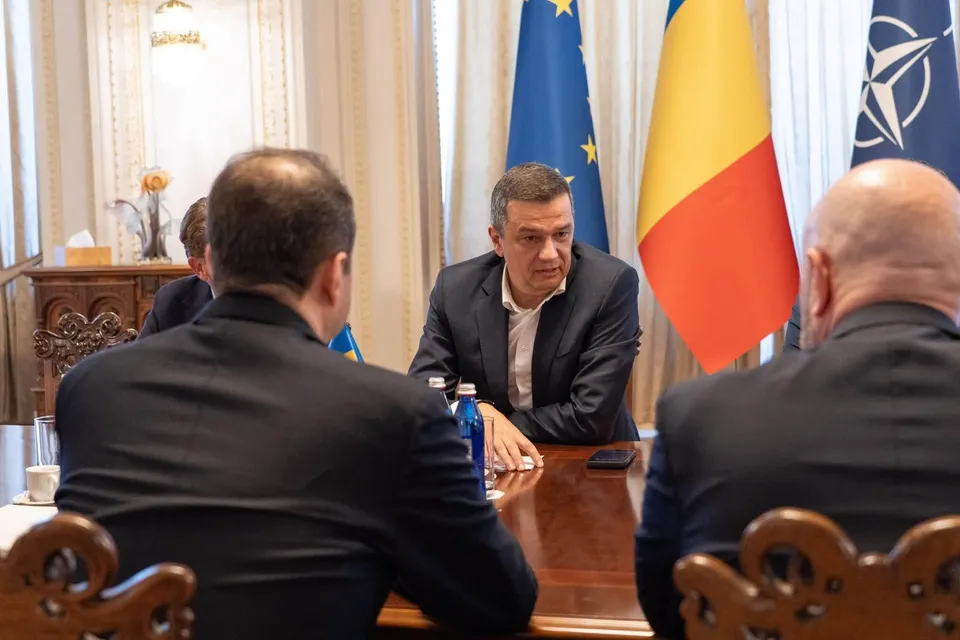 Un bărbat într-un sacou închis la culoare stă la o masă de lemn lângă steaguri, înconjurat de alți trei bărbați văzuți din spate.