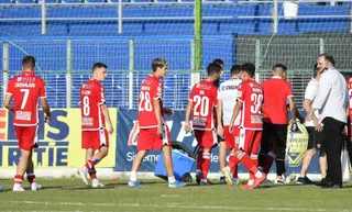 Cei mai mulți jucători de fotbal poartă echipamente roșii și merg pe un teren de fotbal verde în fața unor tribune albastre.