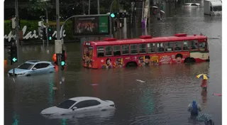Un autobuz roșu și mai multe mașini sunt parțial scufundate în apă pe un drum inundat, lângă semafoare și un panou publicitar.