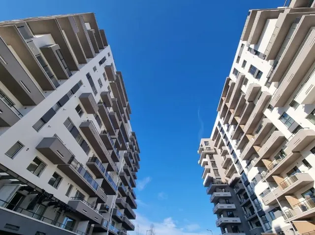 Imaginea arată două blocuri moderne cu balcoane, separate de un cer albastru, văzute dintr-un unghi inferior.