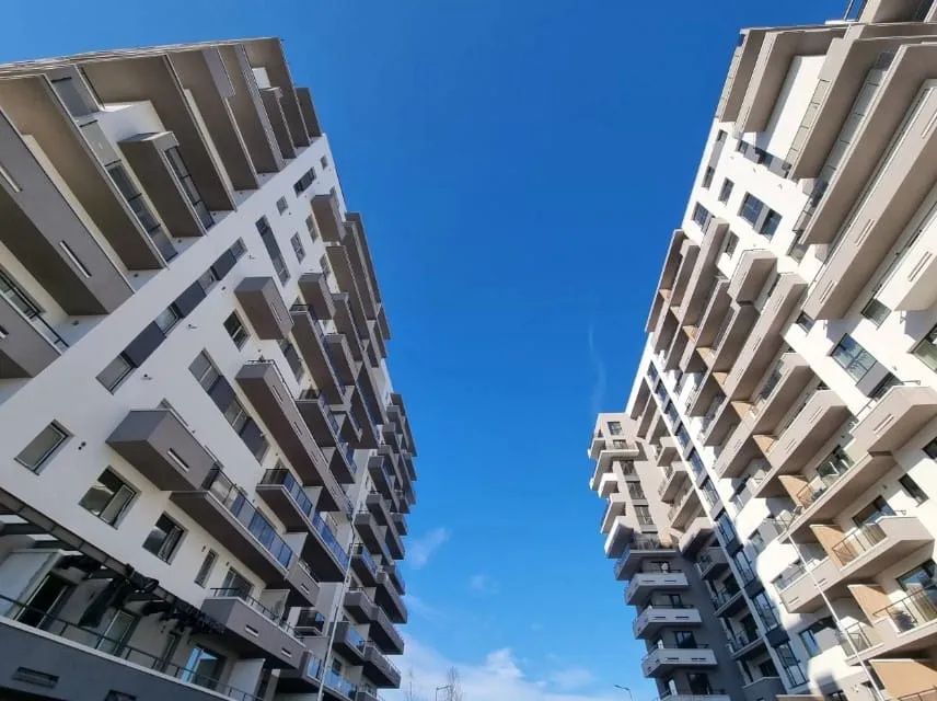 Imaginea arată două blocuri moderne cu balcoane, separate de un cer albastru, văzute dintr-un unghi inferior.
