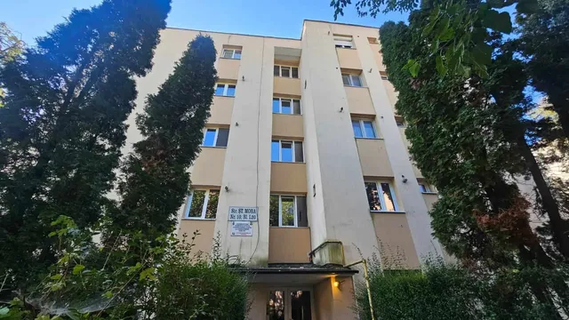 O clădire de apartamente de culoare deschisă este încadrată de copaci verzi și vegetație înaltă sub un cer albastru.
