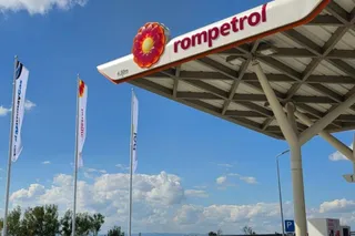 O stație de alimentare Rompetrol sub un cer albastru cu nori albi, având trei steaguri verticale și o structură de acoperiș metalică.