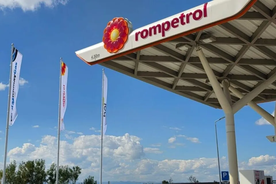 O stație de alimentare Rompetrol sub un cer albastru cu nori albi, având trei steaguri verticale și o structură de acoperiș metalică.