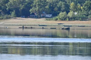 Un râu cu apă calmă reflectă copacii de pe malul opus, unde se află o mașină, o rulotă și vegetație densă.
