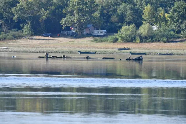 Un râu cu apă calmă reflectă copacii de pe malul opus, unde se află o mașină, o rulotă și vegetație densă.