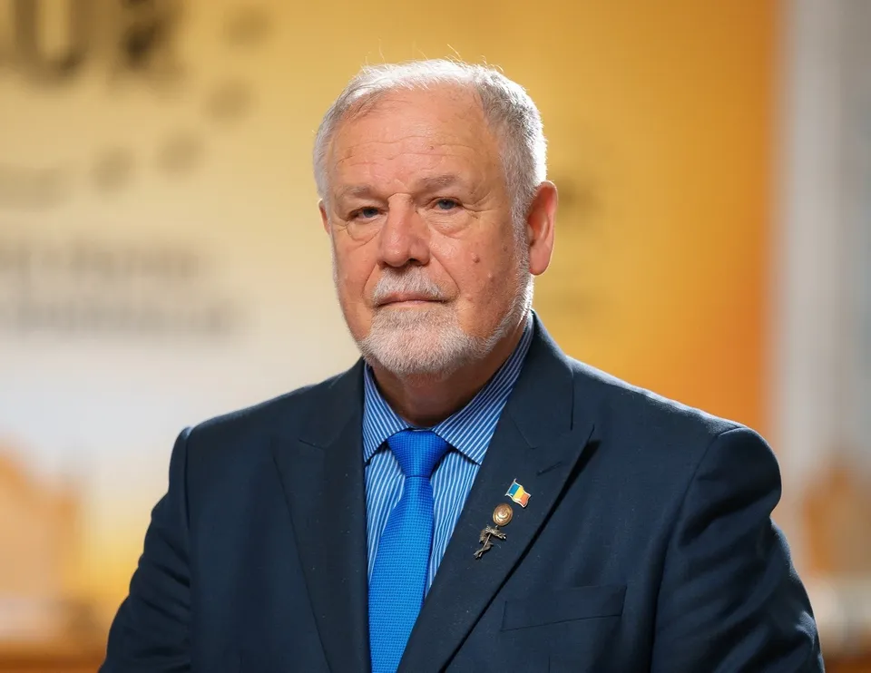 Un bărbat în vârstă, cu barbă și păr alb, poartă un costum bleumarin, cămașă albastră cu dungi și cravată albastră, având o insignă la rever.