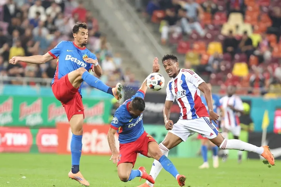 Câțiva jucători de fotbal în echipamente albastre și roșii și unul în echipament alb cu dungi roșii dispută un meci pe un teren verde.
