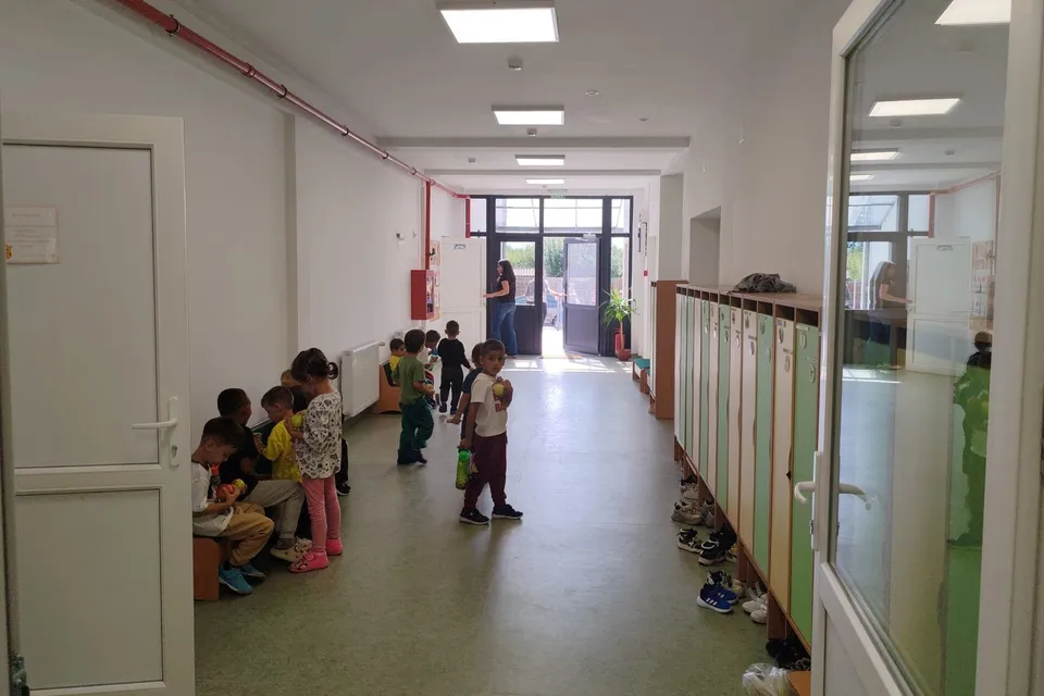 Copii mici stau în grup sau merg pe un coridor interior cu pereți albi și uși de sticlă.