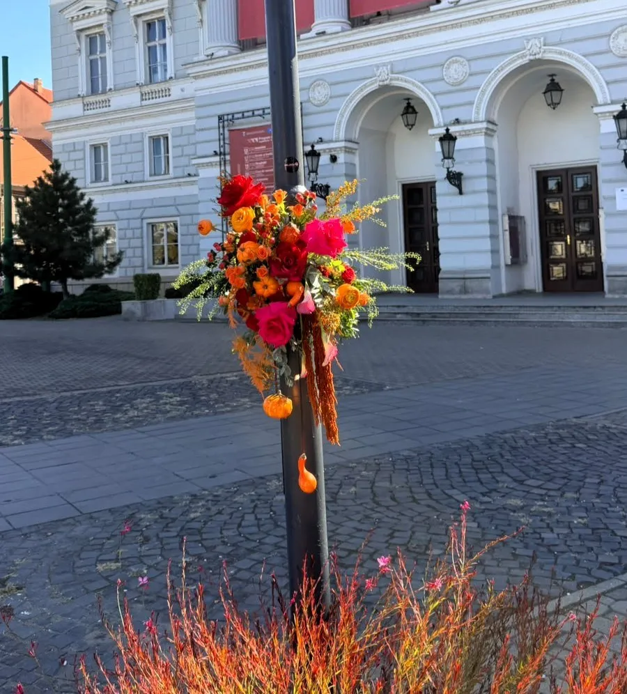 Un aranjament floral colorat cu flori roșii, portocalii și frunze verzi este atașat de un stâlp metalic negru pe o stradă pavată.