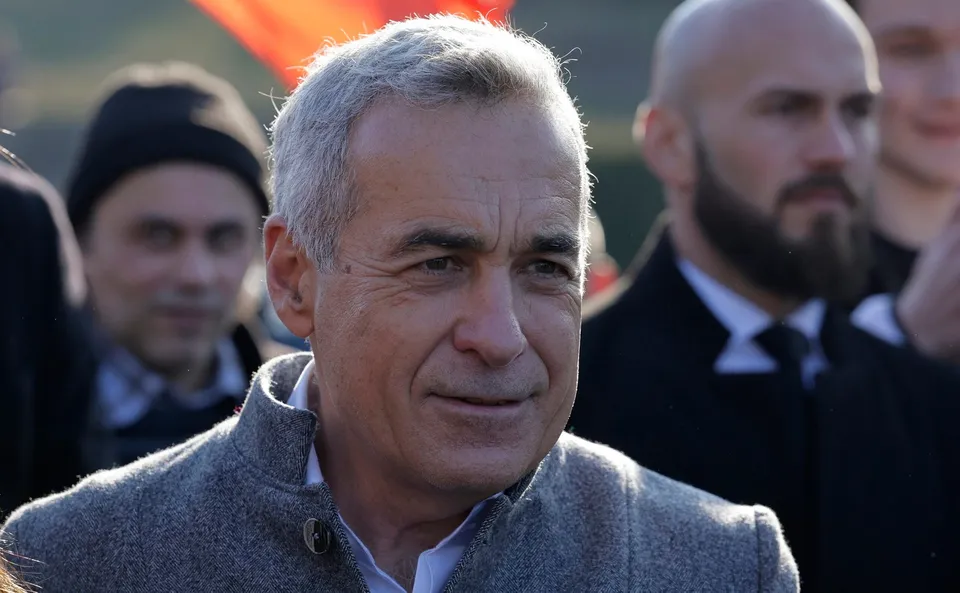 Un bărbat cu părul cărunt și sacou gri este în prim-plan, fiind înconjurat de alți bărbați în planul secund.