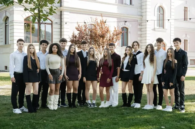 Un grup de tineri stă în rând pe un covor de iarbă verde în fața unei clădiri albe cu ferestre înalte.