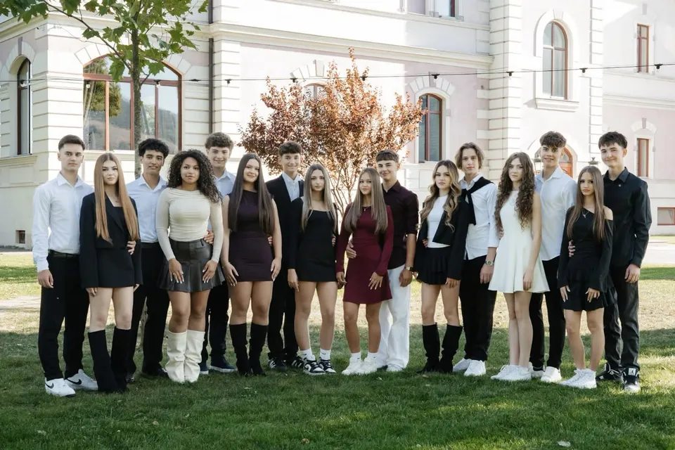 Un grup de tineri stă în rând pe un covor de iarbă verde în fața unei clădiri albe cu ferestre înalte.