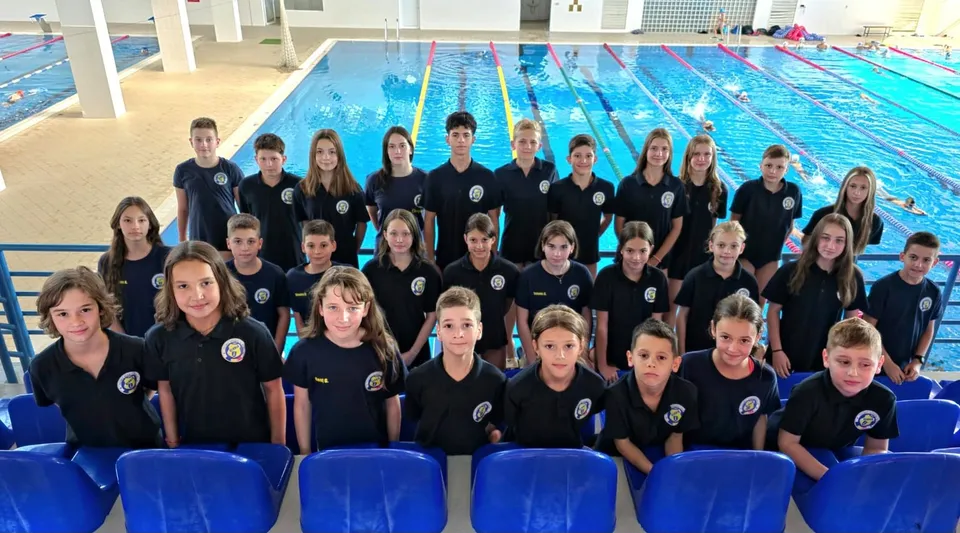 Un grup de copii îmbrăcați în tricouri negre stă așezat în rânduri în fața unei piscine cu apă albastră.