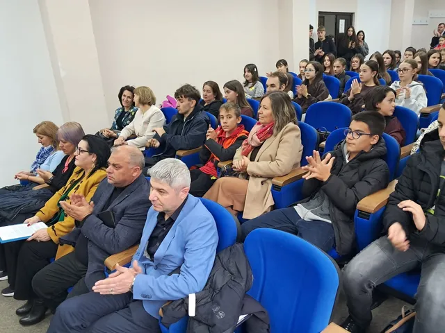 Oamenii stau așezați pe scaune albastre într-o sală, privind spre o direcție comună și aplaudând.