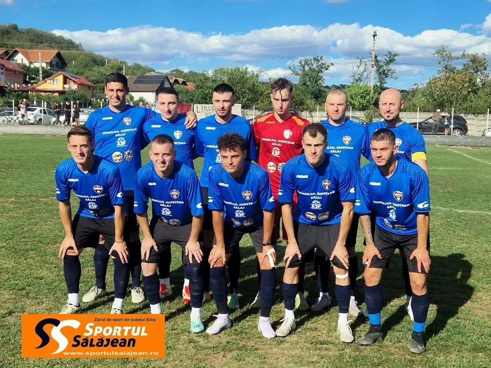 O echipă de fotbaliști în echipament albastru pozează pe un teren de fotbal sub un cer cu nori albe.