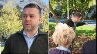 Un bărbat cu barbă și un pulover gri stă în fața unor copaci, în timp ce un alt bărbat arată spre o zonă cu iarbă verde.