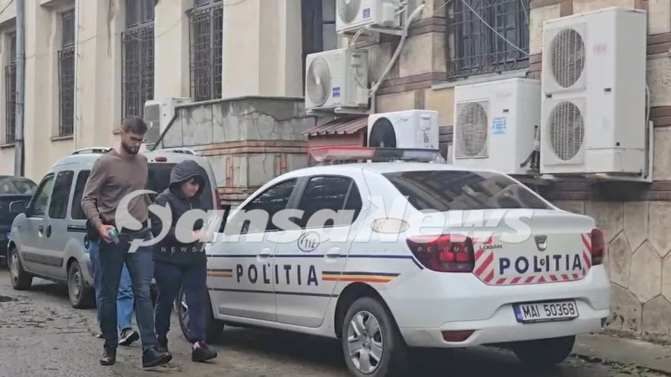 Un bărbat și o femeie stau lângă un vehicul de poliție alb parcat pe o stradă lângă o clădire cu unități de aer condiționat.
