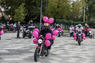 Un motociclist cu cască neagră și tricou negru conduce o motocicletă decorată cu baloane roz pe o suprafață pavată cu plăci gri.