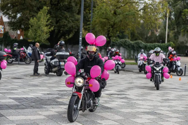 Un motociclist cu cască neagră și tricou negru conduce o motocicletă decorată cu baloane roz pe o suprafață pavată cu plăci gri.