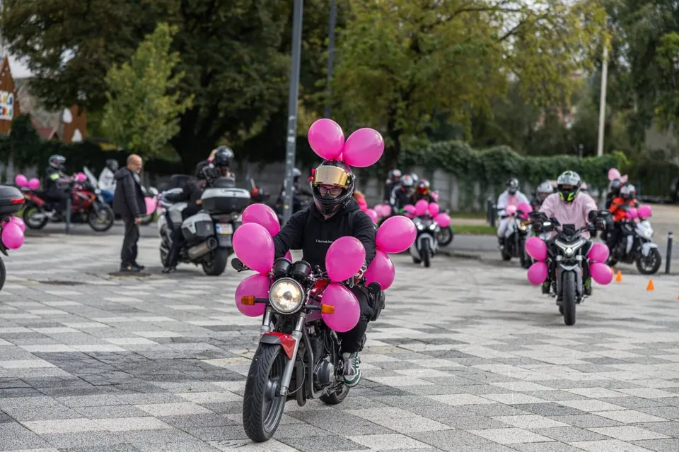 Un motociclist cu cască neagră și tricou negru conduce o motocicletă decorată cu baloane roz pe o suprafață pavată cu plăci gri.