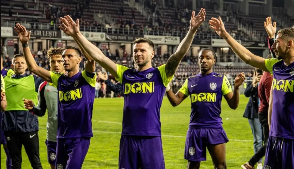 Câțiva bărbați în echipamente sportive mov și galben stau pe un teren de fotbal cu brațele ridicate spre tribunele unui stadion plin.