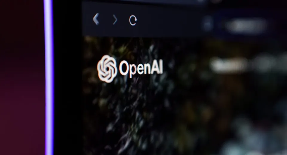 Logoul și textul OpenAI sunt afișate pe un ecran de computer în fața unui fundal întunecat și neclar.