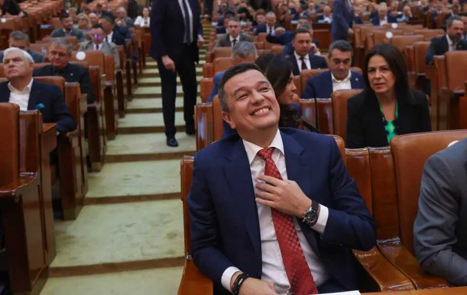 Un bărbat într-un costum albastru și cravată roșie zâmbește, stând într-o sală cu scaune de lemn și mulți oameni așezați în fundal.