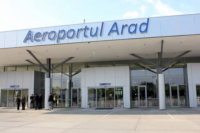 O clădire modernă de aeroport cu inscripția Aeroportul Arad, având suprafețe vitrate și câțiva oameni stând pe trotuarul din fața intrării.