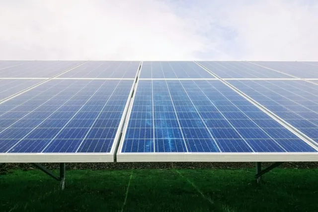 Rânduri de panouri solare albastre sunt instalate pe un suport metalic deasupra unui câmp de iarbă verde sub un cer înnorat.