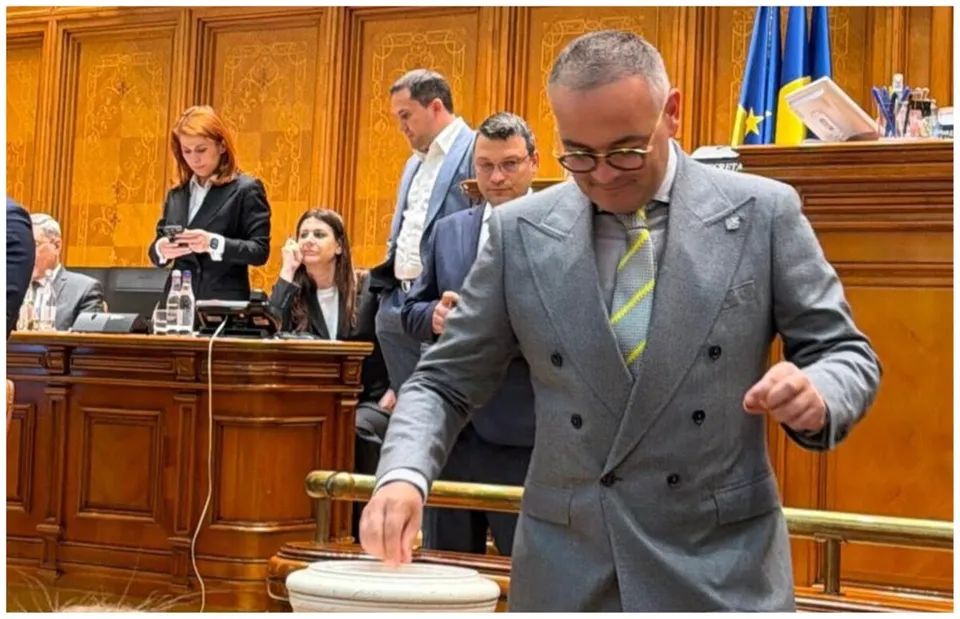 Un bărbat cu ochelari și sacou gri stă în prim-plan, în timp ce alți oameni se află în spatele unui birou de lemn.