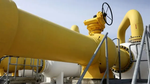 Imaginea prezintă o instalație industrială cu conducte groase, galbene și albe, și o valvă cu roată, sub un cer senin.