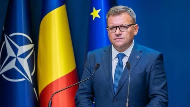 Un bărbat cu ochelari și costum albastru stă în fața unor steaguri, având două microfoane în prim-plan.