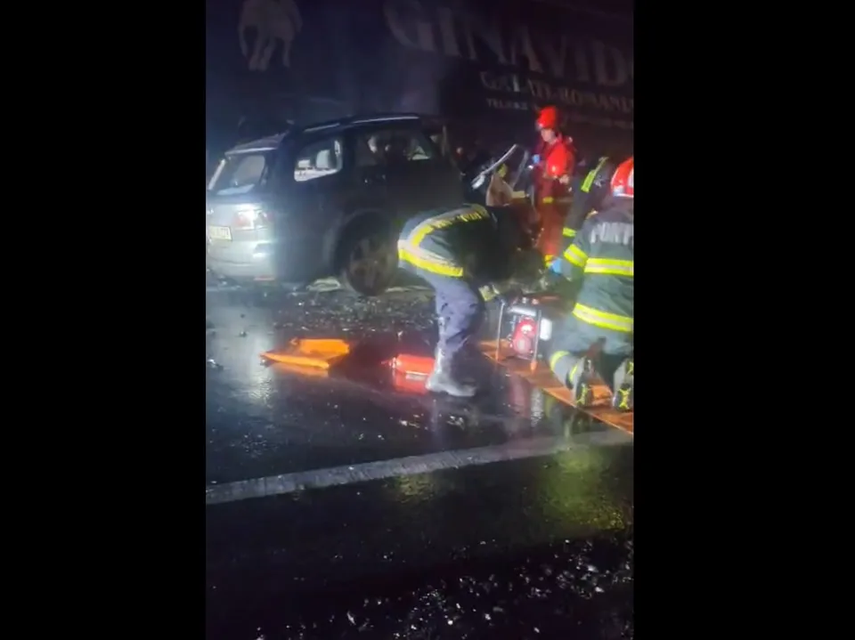 Pompieri în echipament reflectorizant lucrează pe un drum umed lângă un autoturism negru în timpul nopții.