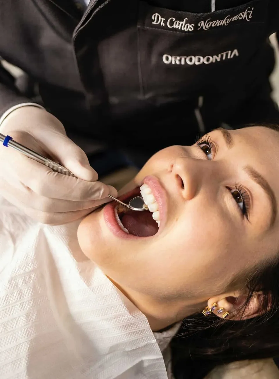 O persoană cu gura deschisă stă întinsă pe un scaun, în timp ce o mână cu mănușă albă folosește o oglindă dentară în cavitatea bucală.