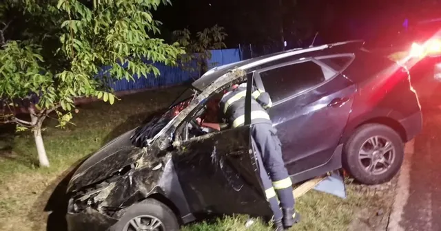 Un om în uniformă de salvator stă lângă un autoturism negru cu partea frontală zdrobită pe marginea unui drum noaptea.
