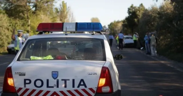 O mașină de poliție albă cu girofaruri colorate este parcată pe un drum, în fața căreia se află alți oameni și vehicule.