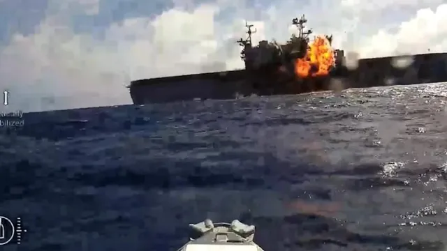O navă mare de culoare închisă navighează pe apă, având o explozie cu flăcări portocalii pe partea sa dreaptă sub un cer înnorat.