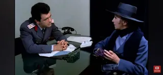 Un bărbat în uniformă gri stă la birou lângă un telefon, privind spre o femeie purtând pălărie și haine albastre.