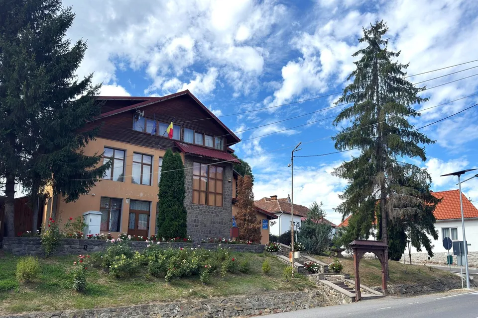 O clădire cu acoperiș înclinat, situată pe un deal cu vegetație, sub un cer cu nori plutitori.