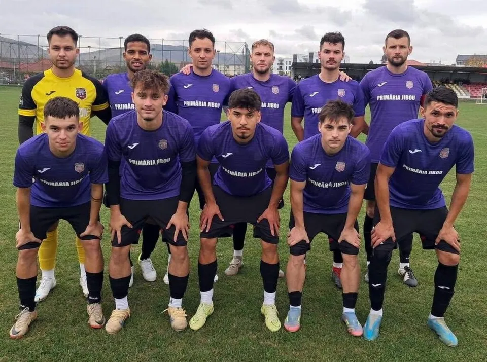 O echipă de fotbal de zece bărbați poartă echipamente sportive de culoare albastră și galbenă, stând pe un teren de iarbă verde.