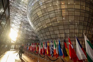 O serie de steaguri colorate este așezată pe un suport în fața unei structuri sferice de sticlă și metal sub lumina soarelui.