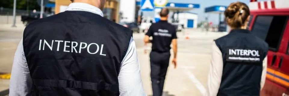 Trei persoane purtând vesta neagră cu inscripția INTERPOL merg pe o stradă lângă un vehicul roșu.