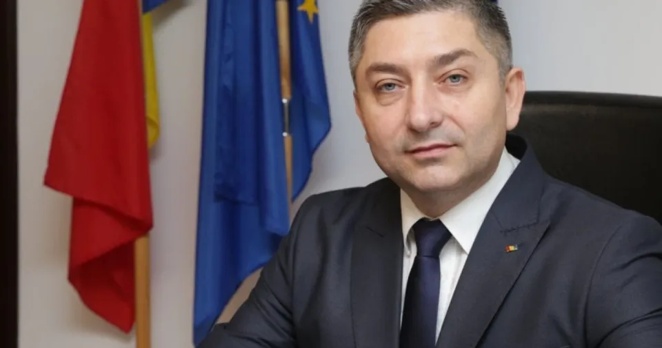 Un bărbat cu părul scurt, purtând costum închis la culoare, cămașă albă și cravată, stă în fața unor steaguri colorate.
